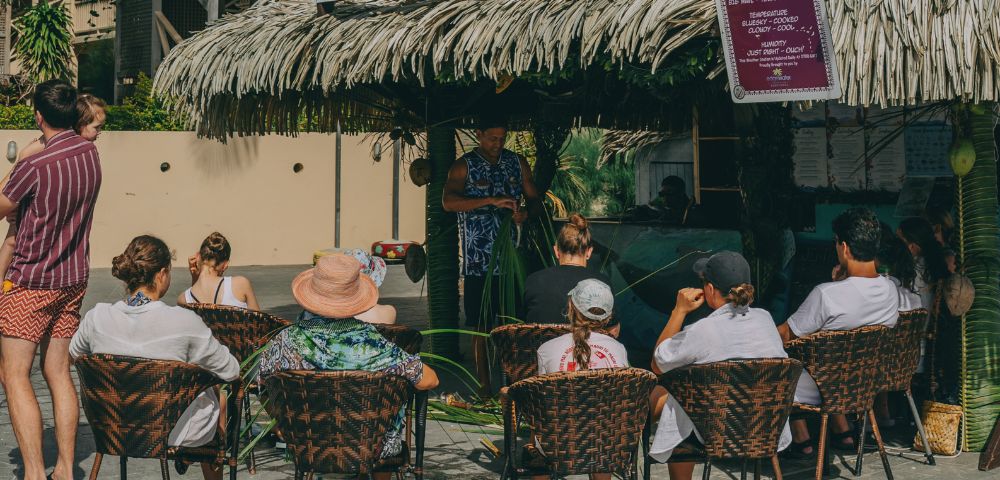A captivating image displays a vibrant scene at a tropical haven, where an image of a thatched hut, bedecked with a striking purple awning, captivates onlookers. Seated on chairs, a lively crowd engages in conversation and feasting, creating a laid-back, inclusive gathering around the focal point.