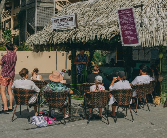 A captivating image displays a vibrant scene at a tropical haven, where an image of a thatched hut, bedecked with a striking purple awning, captivates onlookers. Seated on chairs, a lively crowd engages in conversation and feasting, creating a laid-back, inclusive gathering around the focal point.