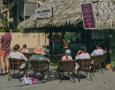 A captivating image displays a vibrant scene at a tropical haven, where an image of a thatched hut, bedecked with a striking purple awning, captivates onlookers. Seated on chairs, a lively crowd engages in conversation and feasting, creating a laid-back, inclusive gathering around the focal point.