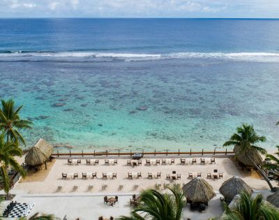 Scenic beachfront with calm blue ocean, sandy shore, palm trees, thatched-roof huts, and rows of lounge chairs under a partly cloudy sky.