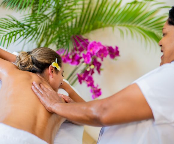 A person receives a back massage from a masseuse in a spa setting, with tropical flowers and greenery in the background.