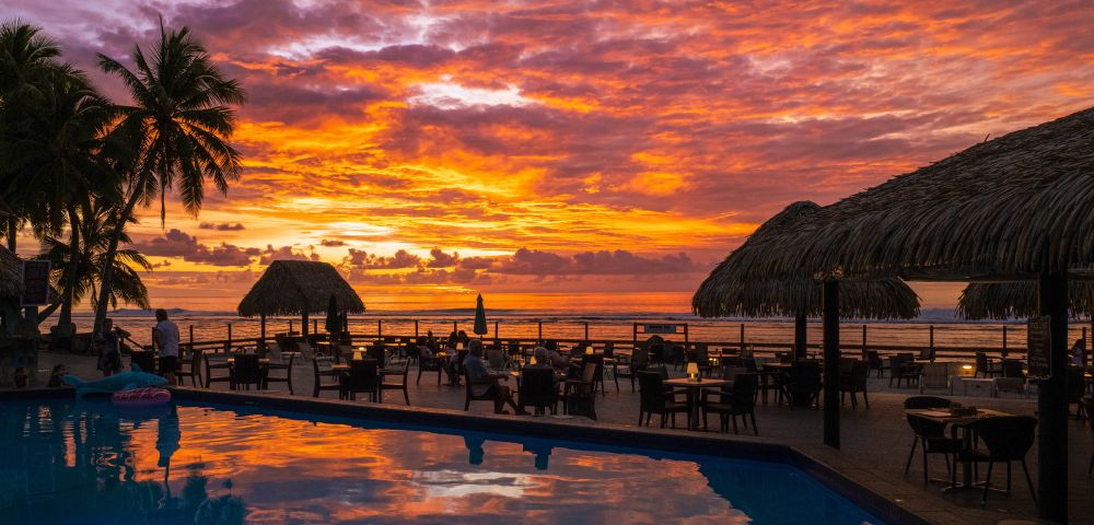 Sunset at a beachside resort with vibrant sky colors, palm trees, thatched-roof structures, a reflective swimming pool, and guests dining outdoors.