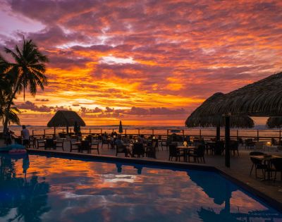 Sunset at a beachside resort with vibrant sky colors, palm trees, thatched-roof structures, a reflective swimming pool, and guests dining outdoors.