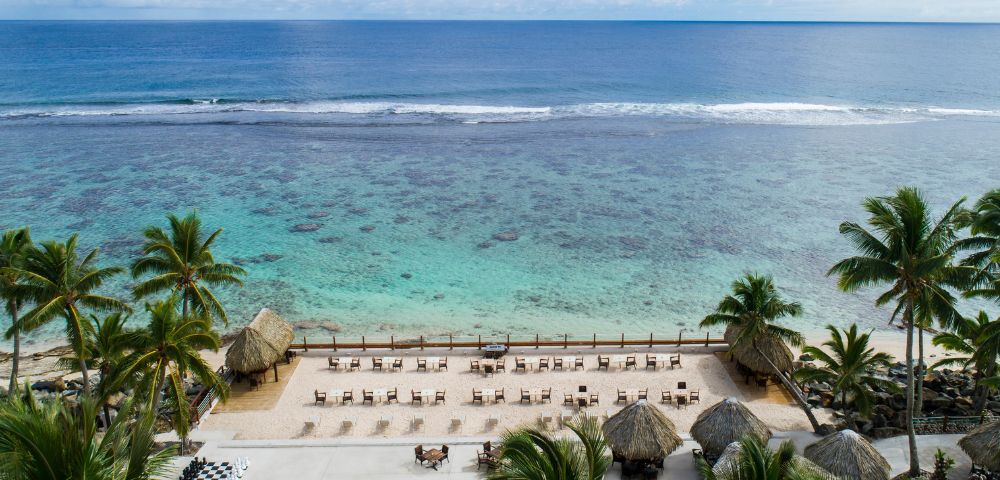 Scenic beachfront with calm blue ocean, sandy shore, palm trees, thatched-roof huts, and rows of lounge chairs under a partly cloudy sky.