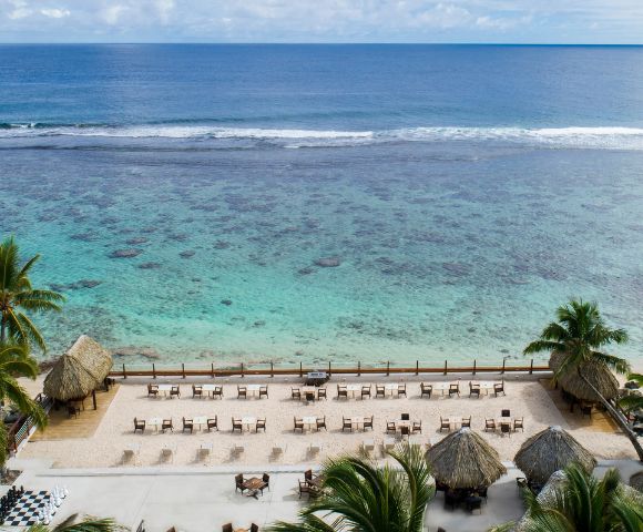 Scenic beachfront with calm blue ocean, sandy shore, palm trees, thatched-roof huts, and rows of lounge chairs under a partly cloudy sky.