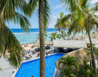 Tropical resort with palm trees, a clear blue swimming pool, thatched-roof structures, and beachfront dining overlooking the ocean.
