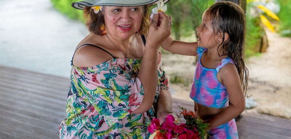 A young girl places a flower in the hat of an older woman wearing an off-the-shoulder floral dress and sun hat, holding a basket of flowers.
