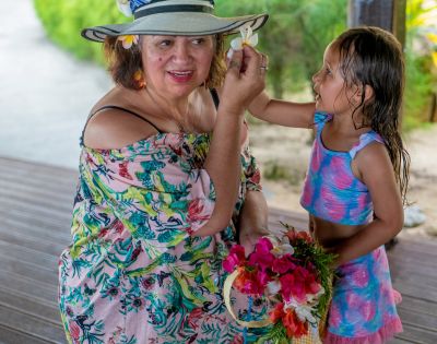 A young girl places a flower in the hat of an older woman wearing an off-the-shoulder floral dress and sun hat, holding a basket of flowers.
