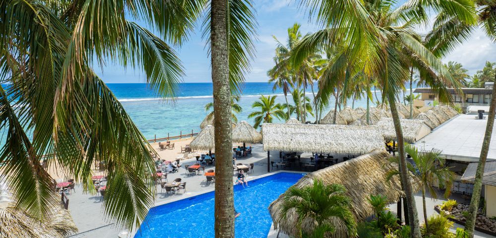 Tropical resort with palm trees, a clear blue swimming pool, thatched-roof structures, and beachfront dining overlooking the ocean.