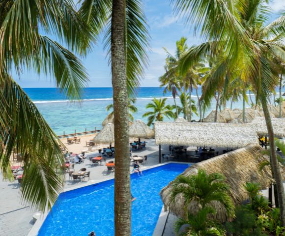 Tropical resort with palm trees, a clear blue swimming pool, thatched-roof structures, and beachfront dining overlooking the ocean.