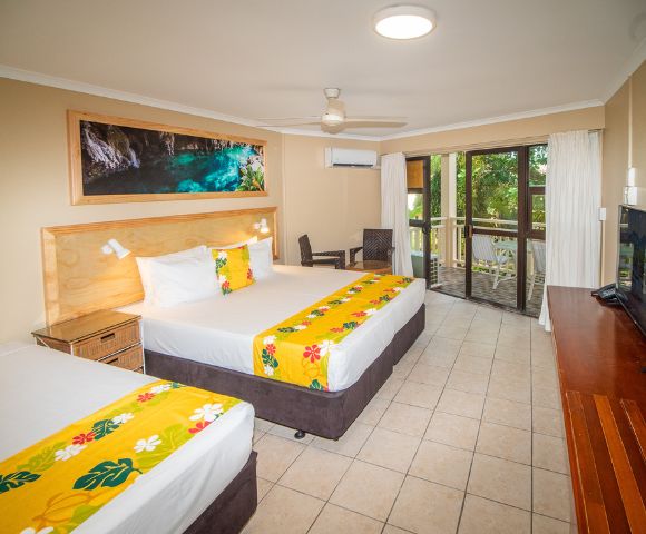Hotel room interior featuring two beds with white linens, yellow floral runners, and matching yellow decorative pillows, a wooden headboard, a large framed art piece above the beds depicting a water cave, a flatscreen TV on a wooden console, and a balcony visible through sliding glass doors.