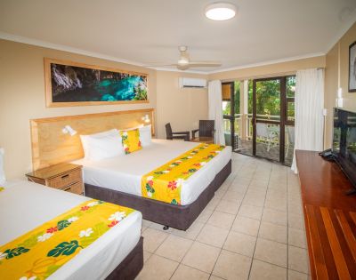 Hotel room interior featuring two beds with white linens, yellow floral runners, and matching yellow decorative pillows, a wooden headboard, a large framed art piece above the beds depicting a water cave, a flatscreen TV on a wooden console, and a balcony visible through sliding glass doors.