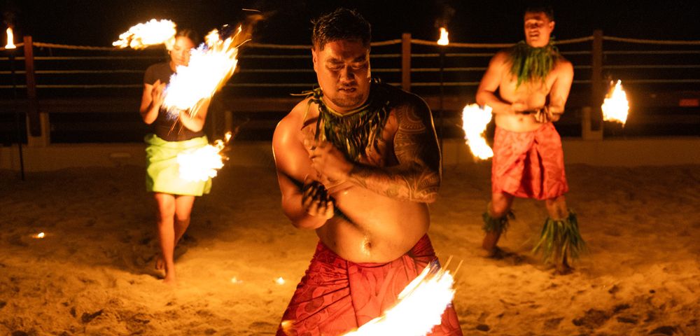 Three fire dancers performing on a sandy beach at night, two men and a woman, each holding a flaming torch. The central man is the focus, with tattoos on his arm, wearing a red wrap and a leaf necklace, moving with a torch held low. Other dancers stand on either side with fire fans and in similar traditional attire, all lit by the torch flames and surrounding torches.
