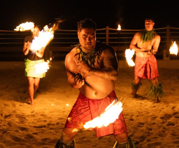 Three fire dancers performing on a sandy beach at night, two men and a woman, each holding a flaming torch. The central man is the focus, with tattoos on his arm, wearing a red wrap and a leaf necklace, moving with a torch held low. Other dancers stand on either side with fire fans and in similar traditional attire, all lit by the torch flames and surrounding torches.