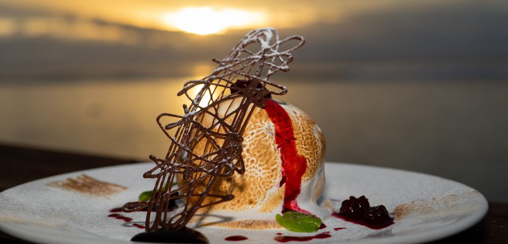 Baked Alaska dessert on a white plate, featuring a dome of ice cream and meringue with a browned exterior, drizzled with red fruit sauce, and garnished with a chocolate lattice and mint sprig, set against a blurred sunset over water.