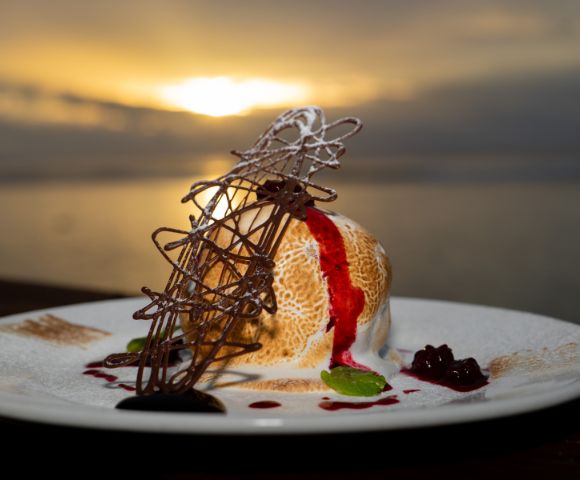 Baked Alaska dessert on a white plate, featuring a dome of ice cream and meringue with a browned exterior, drizzled with red fruit sauce, and garnished with a chocolate lattice and mint sprig, set against a blurred sunset over water.
