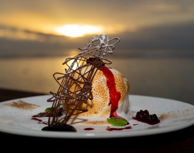 Baked Alaska dessert on a white plate, featuring a dome of ice cream and meringue with a browned exterior, drizzled with red fruit sauce, and garnished with a chocolate lattice and mint sprig, set against a blurred sunset over water.