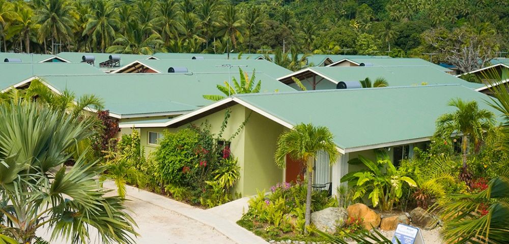 Overhead shot of a tropical resort with multiple single-story buildings with green roofs and light green exteriors, surrounded by lush greenery, palm trees, and tropical plants, nestled against a hillside covered in dense forest.