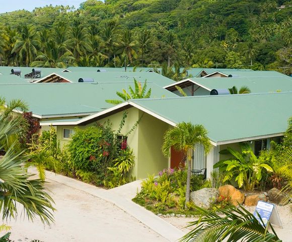 Overhead shot of a tropical resort with multiple single-story buildings with green roofs and light green exteriors, surrounded by lush greenery, palm trees, and tropical plants, nestled against a hillside covered in dense forest.