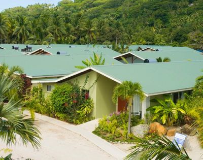 Overhead shot of a tropical resort with multiple single-story buildings with green roofs and light green exteriors, surrounded by lush greenery, palm trees, and tropical plants, nestled against a hillside covered in dense forest.