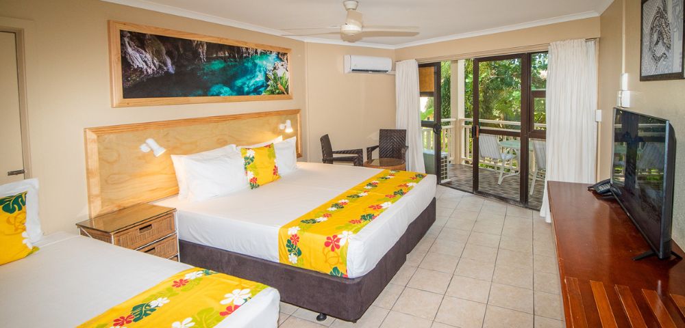 Hotel room interior featuring two beds with white linens, yellow floral runners, and matching yellow decorative pillows, a wooden headboard, a large framed art piece above the beds depicting a water cave, a flatscreen TV on a wooden console, and a balcony visible through sliding glass doors.