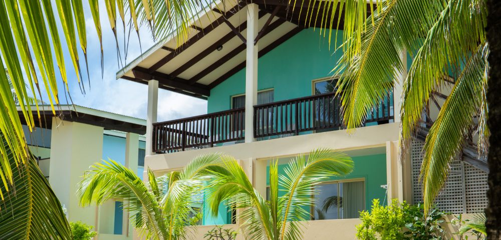 Two-story tropical building with turquoise exterior and dark brown trim, accented by palm fronds in the foreground, suggesting a resort setting.