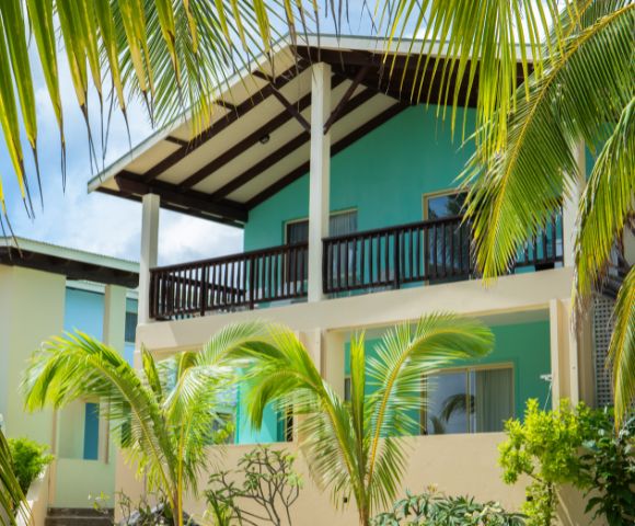 Two-story tropical building with turquoise exterior and dark brown trim, accented by palm fronds in the foreground, suggesting a resort setting.