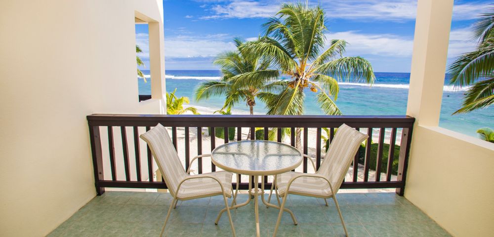 Balcony overlooking a tropical beach scene; two beige chairs flank a round glass table, brown wooden railing, open to a sandy beach with palm trees, and a turquoise ocean under a bright blue sky.