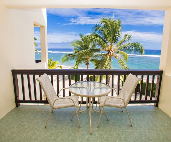 Balcony overlooking a tropical beach scene; two beige chairs flank a round glass table, brown wooden railing, open to a sandy beach with palm trees, and a turquoise ocean under a bright blue sky.