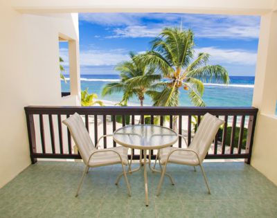 Balcony overlooking a tropical beach scene; two beige chairs flank a round glass table, brown wooden railing, open to a sandy beach with palm trees, and a turquoise ocean under a bright blue sky.