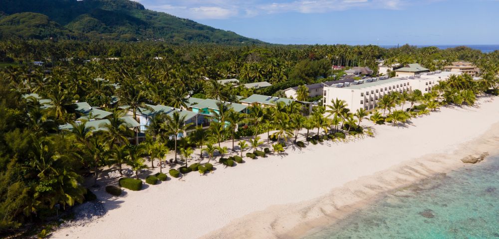 Aerial view of a tropical resort on a white sand beach, featuring low-rise buildings with green roofs, a large multi-story hotel building, palm trees, and turquoise water along a coastline with a backdrop of lush green mountains.