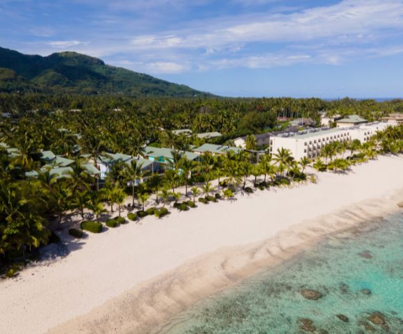 Aerial view of a tropical resort on a white sand beach, featuring low-rise buildings with green roofs, a large multi-story hotel building, palm trees, and turquoise water along a coastline with a backdrop of lush green mountains.