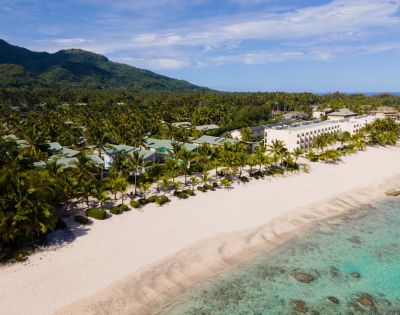 Aerial view of a tropical resort on a white sand beach, featuring low-rise buildings with green roofs, a large multi-story hotel building, palm trees, and turquoise water along a coastline with a backdrop of lush green mountains.