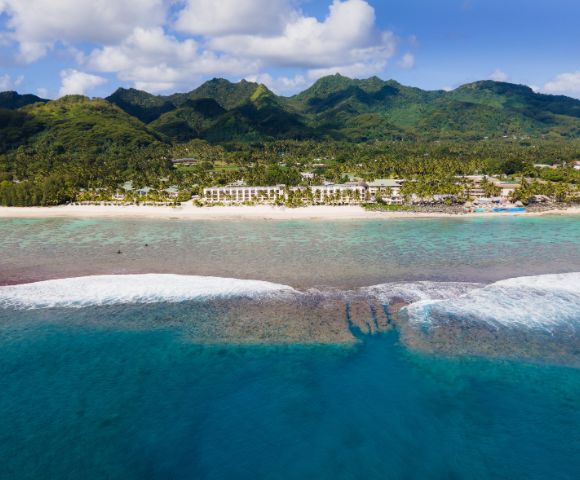 Aerial view of a tropical coastline with a white sand beach, resorts, and lush green mountains, turquoise waters with coral reefs, and waves crashing on the shallow reef.