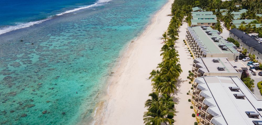 Aerial view of a serene beach with turquoise waters, coral reefs, and palm trees lining beachfront resort buildings.