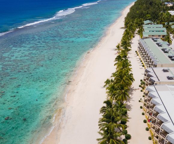 Aerial view of a serene beach with turquoise waters, coral reefs, and palm trees lining beachfront resort buildings.