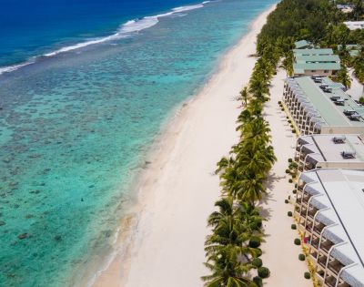 Aerial view of a serene beach with turquoise waters, coral reefs, and palm trees lining beachfront resort buildings.