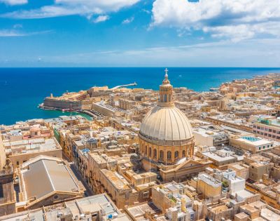 Aerial view of a coastal city with a prominent domed cathedral and a spire, surrounded by rustic buildings. The blue sea and sky add a tranquil backdrop.