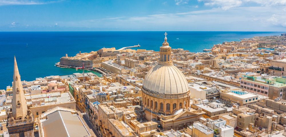 Aerial view of a coastal city with a prominent domed cathedral and a spire, surrounded by rustic buildings. The blue sea and sky add a tranquil backdrop.