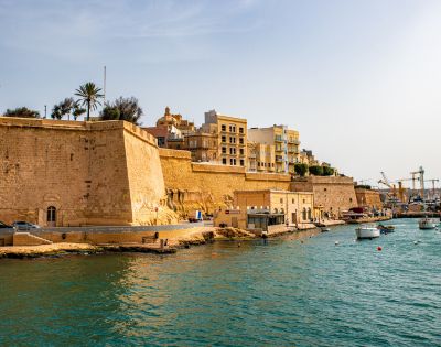 Historic fort with tall walls on a sunny day, against clear blue skies. Boats float in teal waters near the shore. Buildings and cranes in the distance.