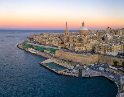 Skyline of Valletta, Malta at sunrise. Prominent dome and spire rise above historic stone buildings. Warm hues illuminate the waterfront, contrasting with the blue sea.