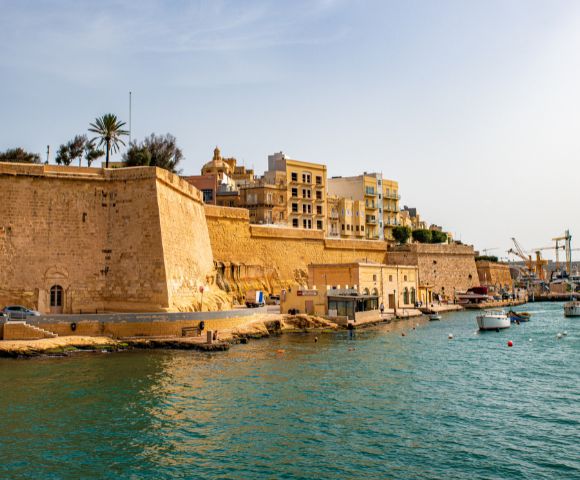 Historic fort with tall walls on a sunny day, against clear blue skies. Boats float in teal waters near the shore. Buildings and cranes in the distance.