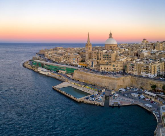 Skyline of Valletta, Malta at sunrise. Prominent dome and spire rise above historic stone buildings. Warm hues illuminate the waterfront, contrasting with the blue sea.