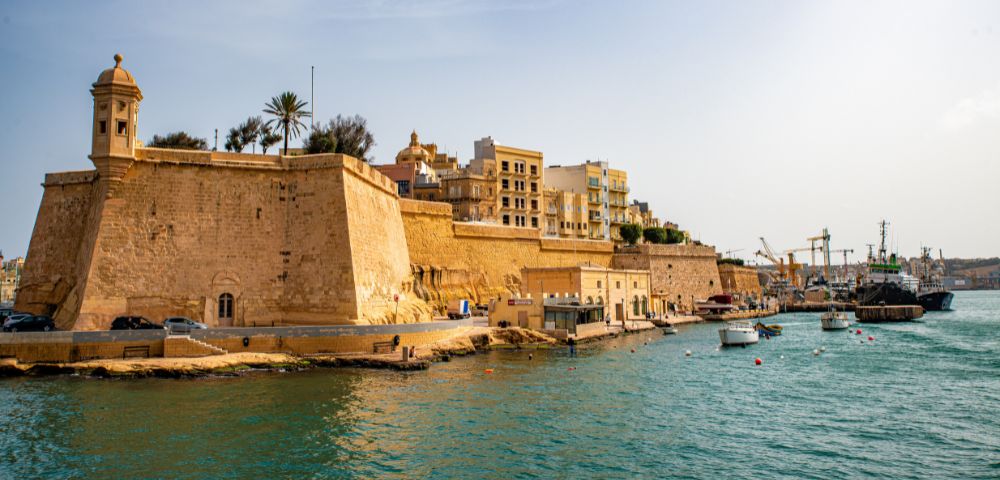 Historic fort with tall walls on a sunny day, against clear blue skies. Boats float in teal waters near the shore. Buildings and cranes in the distance.