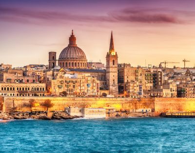 Aerial view of a coastal city at dusk, featuring historic buildings with a prominent dome and spire. The calm sea borders the city, under a pink-orange sky.