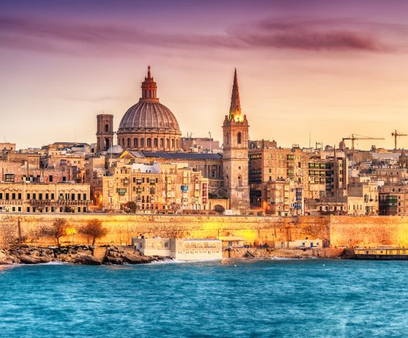 Aerial view of a coastal city at dusk, featuring historic buildings with a prominent dome and spire. The calm sea borders the city, under a pink-orange sky.