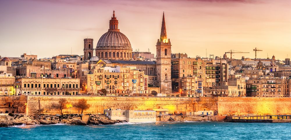 Aerial view of a coastal city at dusk, featuring historic buildings with a prominent dome and spire. The calm sea borders the city, under a pink-orange sky.