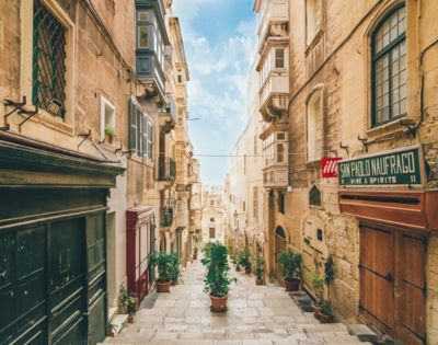 Narrow cobblestone street in a historic European city, lined with tan stone buildings and potted plants. A clear blue sky enhances the picturesque scene.