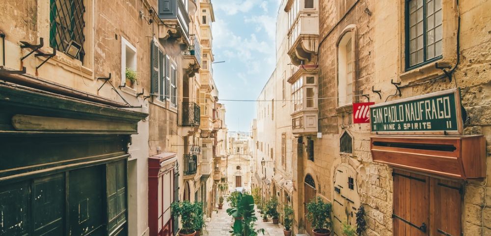 Narrow cobblestone street in a historic European city, lined with tan stone buildings and potted plants. A clear blue sky enhances the picturesque scene.