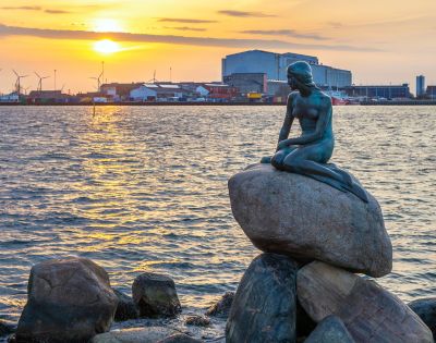 Statue of a mermaid sits on rocks, gazing over a harbor at sunset. Wind turbines and industrial buildings line the distant horizon. Peaceful ambiance.